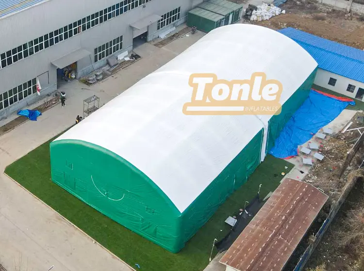 Aerial view of a large white and green inflatable tent with an arched roof situated on a grassy patch near an industrial building.